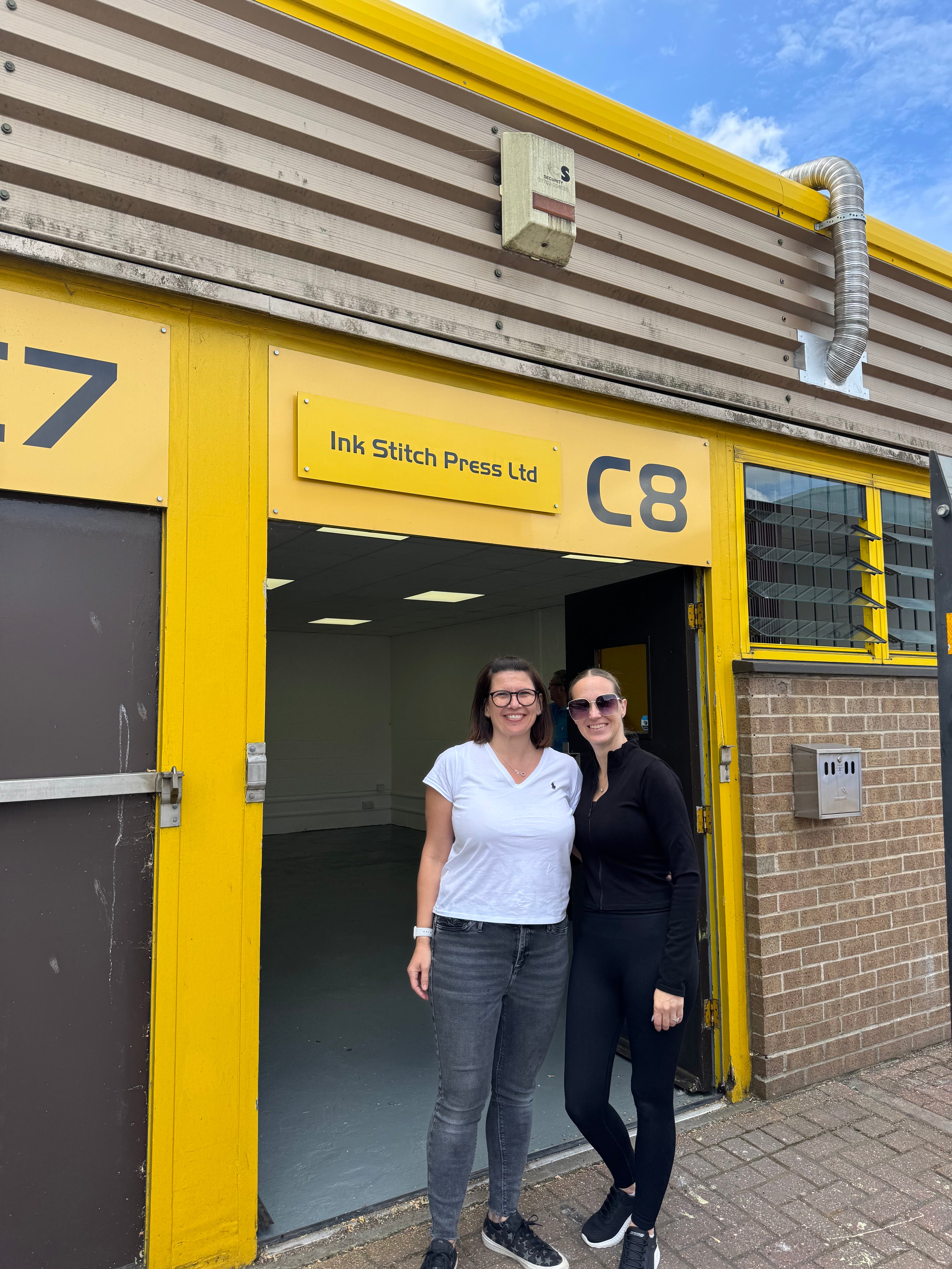 Kelly and Louise, the team behind Ink Stitch Press, posing outside their new yellow and black premises on moving day, marking an exciting milestone for the business.