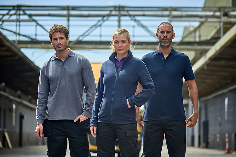 Three workers showcasing RTX workwear in an industrial outdoor setting, featuring a fleece jacket and polo shirts in navy blue and grey, highlighting the brand's versatile and durable clothing options.