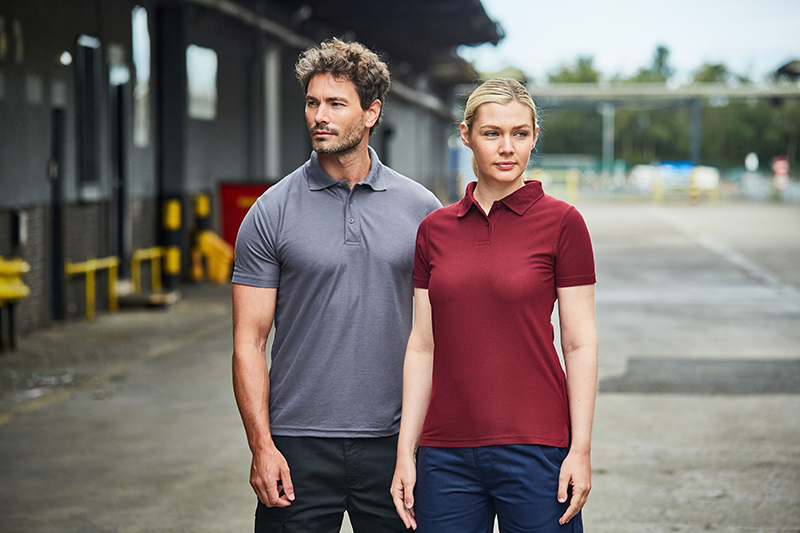 Two workers showcasing RTX workwear polo shirts, one in maroon and the other in grey, standing together in an industrial outdoor setting, highlighting the professional and durable design of the apparel.